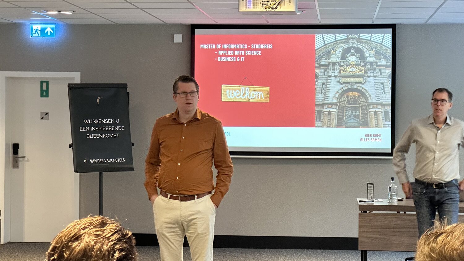 Two hosts stand in front of a red welcome slide reading Master of Informatics studiereis, Applied Data Science, Business and IT, with a photograph of Antwerp Central Station.