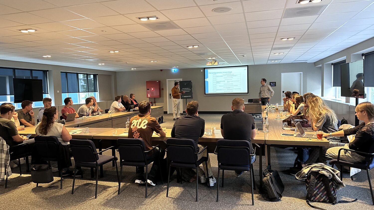 The full room seen from the back, delegates seated around the U-shaped table facing the presenter at the far end.