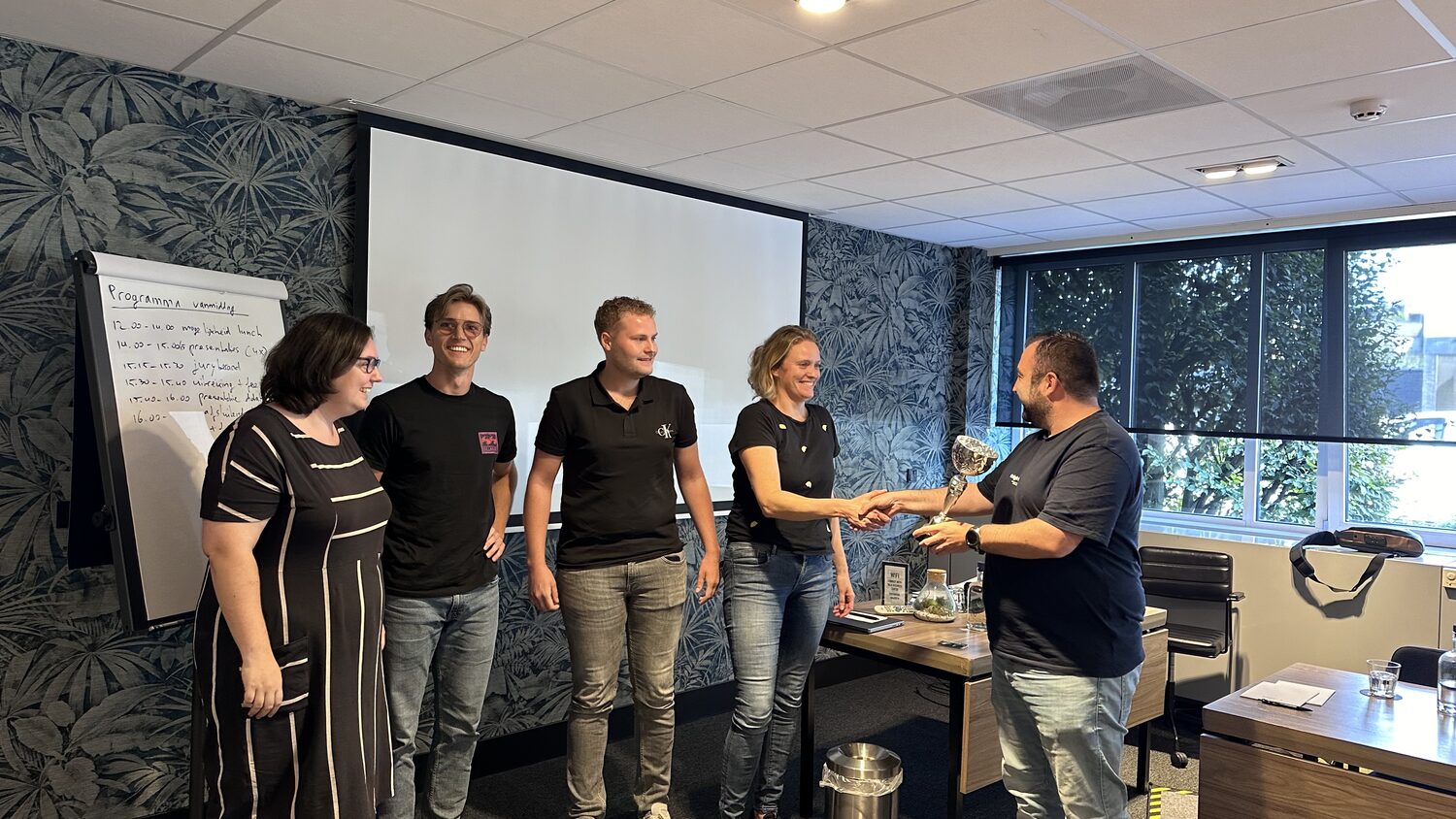 A trophy is handed over with a handshake as the team stands in line beside the programme flip chart.