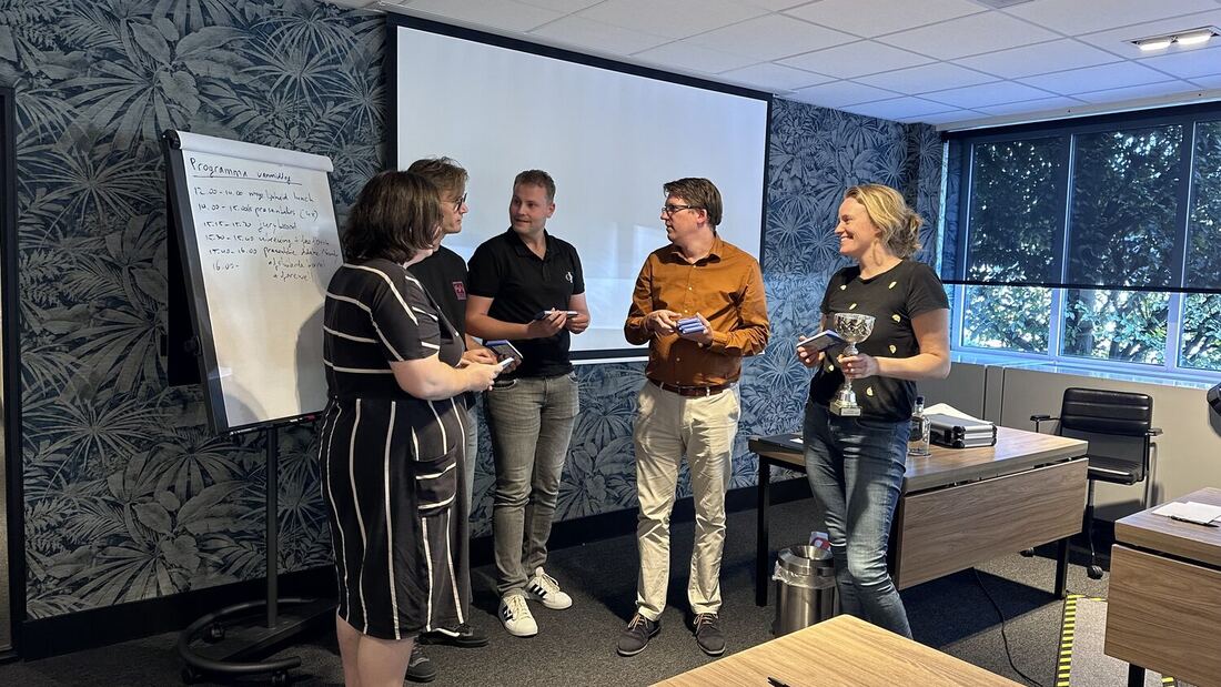Four people hold trophies at the front of the room beside the programme flip chart.