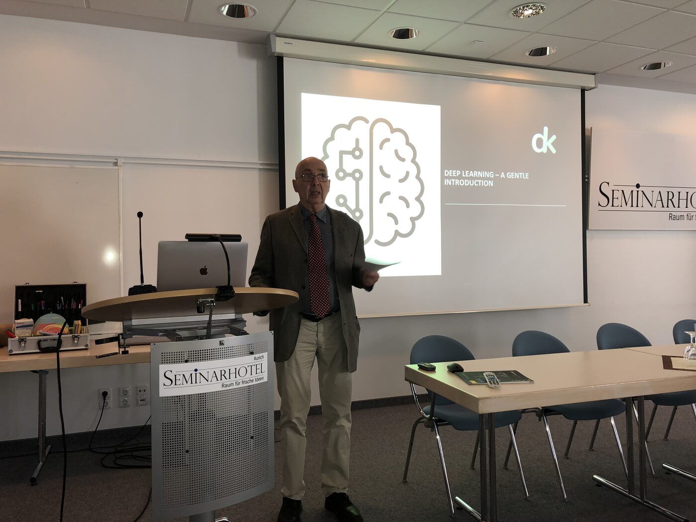 A speaker addresses the room from the lectern at Seminarhotel Aurich. The screen behind carries the deepkapha title slide, a line-drawn brain with circuitry and the words Deep Learning: A Gentle Introduction.
