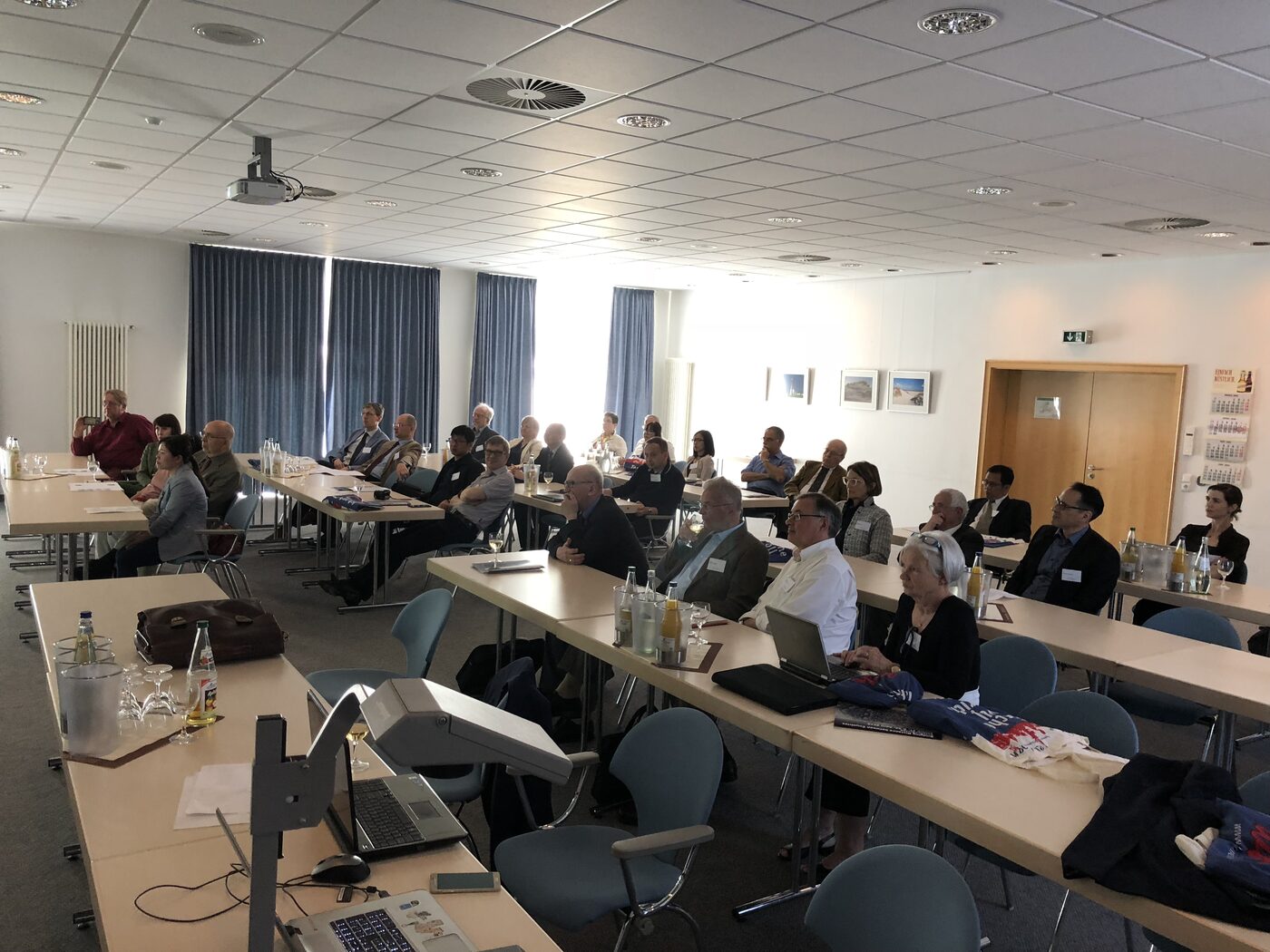 The full meeting room during a session, around thirty delegates in name badges at tables facing the screen, laptops and notes in front of them.