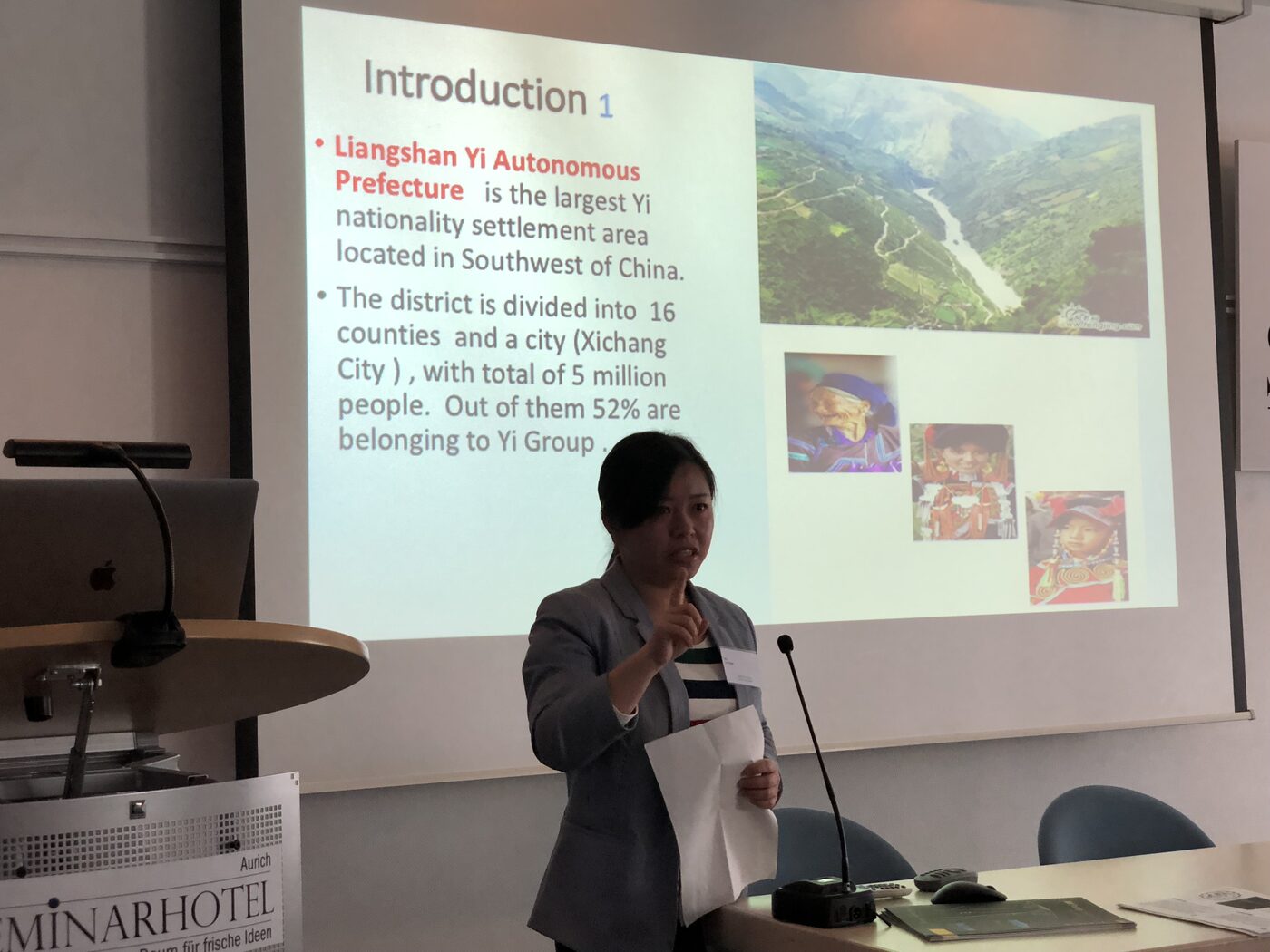 Chen Li presents at the lectern, gesturing to her slide on Liangshan Yi Autonomous Prefecture in southwest China and its sixteen counties and one city.