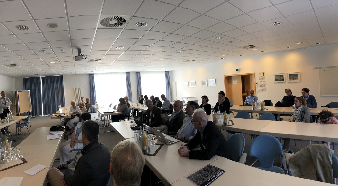 The meeting room from the back, delegates seated around three sides of the table arrangement, the programme booklet for Bridging Distance between Countries open on the table in the foreground.
