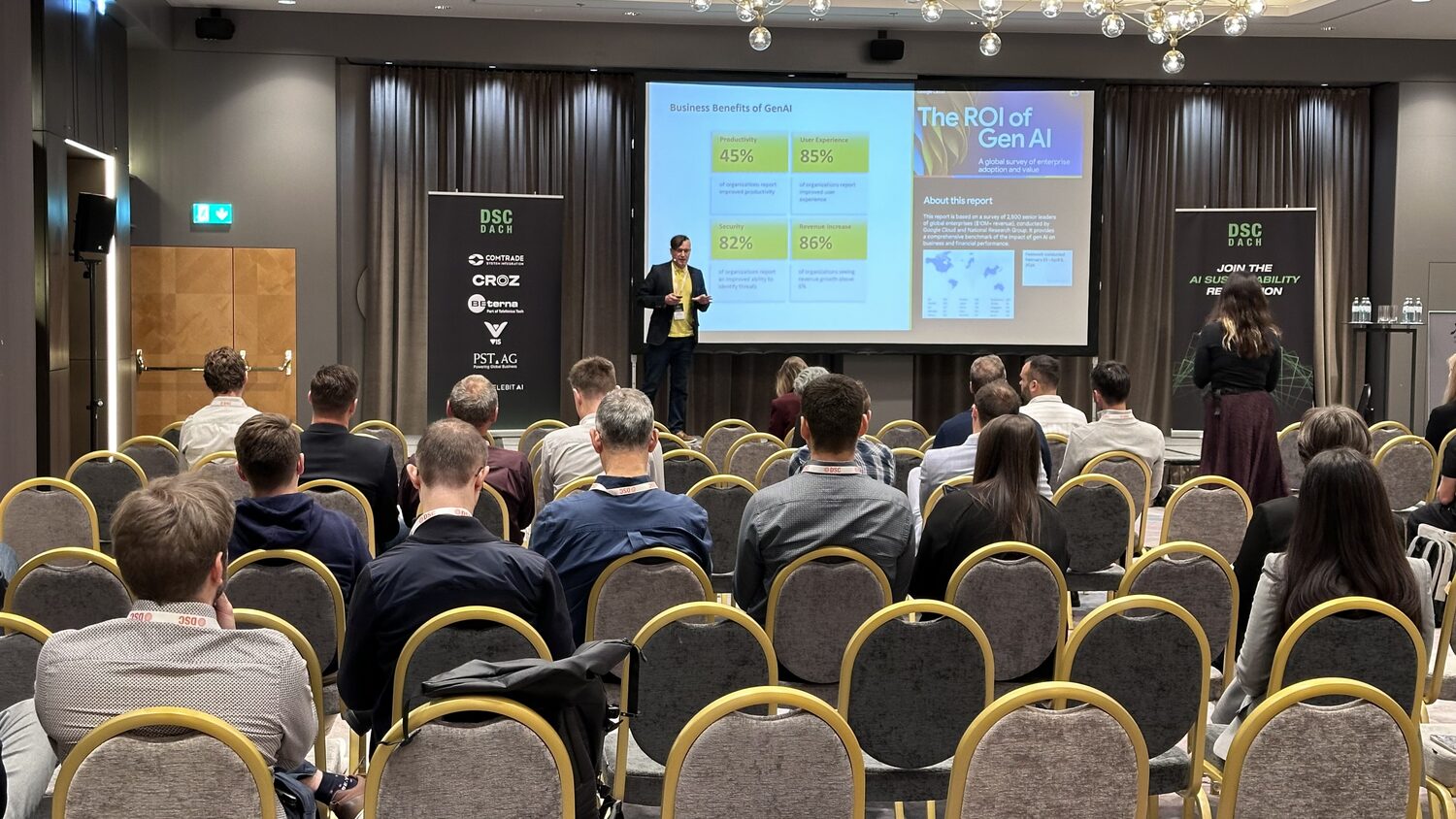 Delegates fill rows of banquet chairs in the main hall while a speaker presents a slide titled The ROI of Gen AI, DSC DACH roll-up banners either side of the screen.