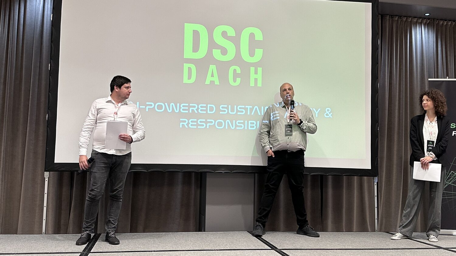 Three people on stage at the opening of DSC DACH, one speaking into a microphone, the screen behind them reading DSC DACH, AI-powered sustainability and responsibility.
