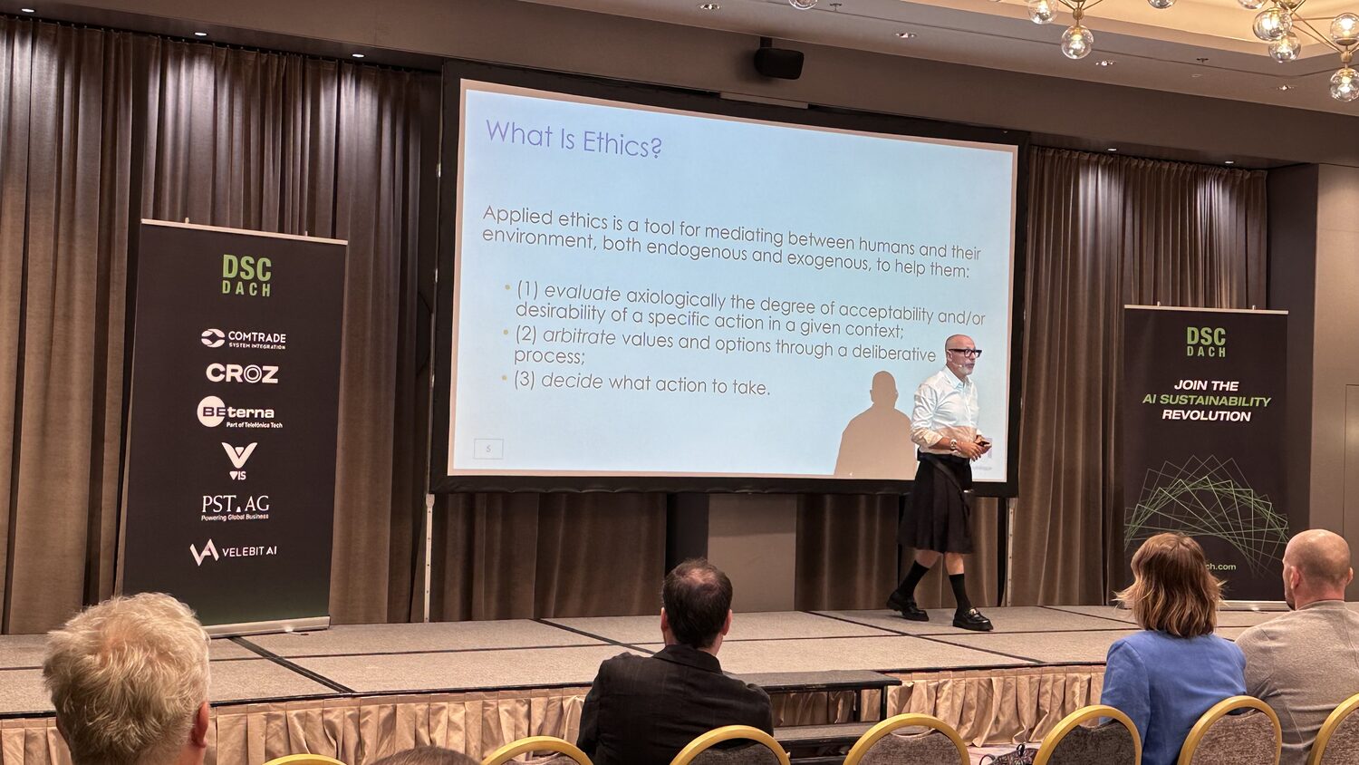 A speaker crosses the stage in front of a slide headed What is Ethics?, setting out a three-part definition of applied ethics.