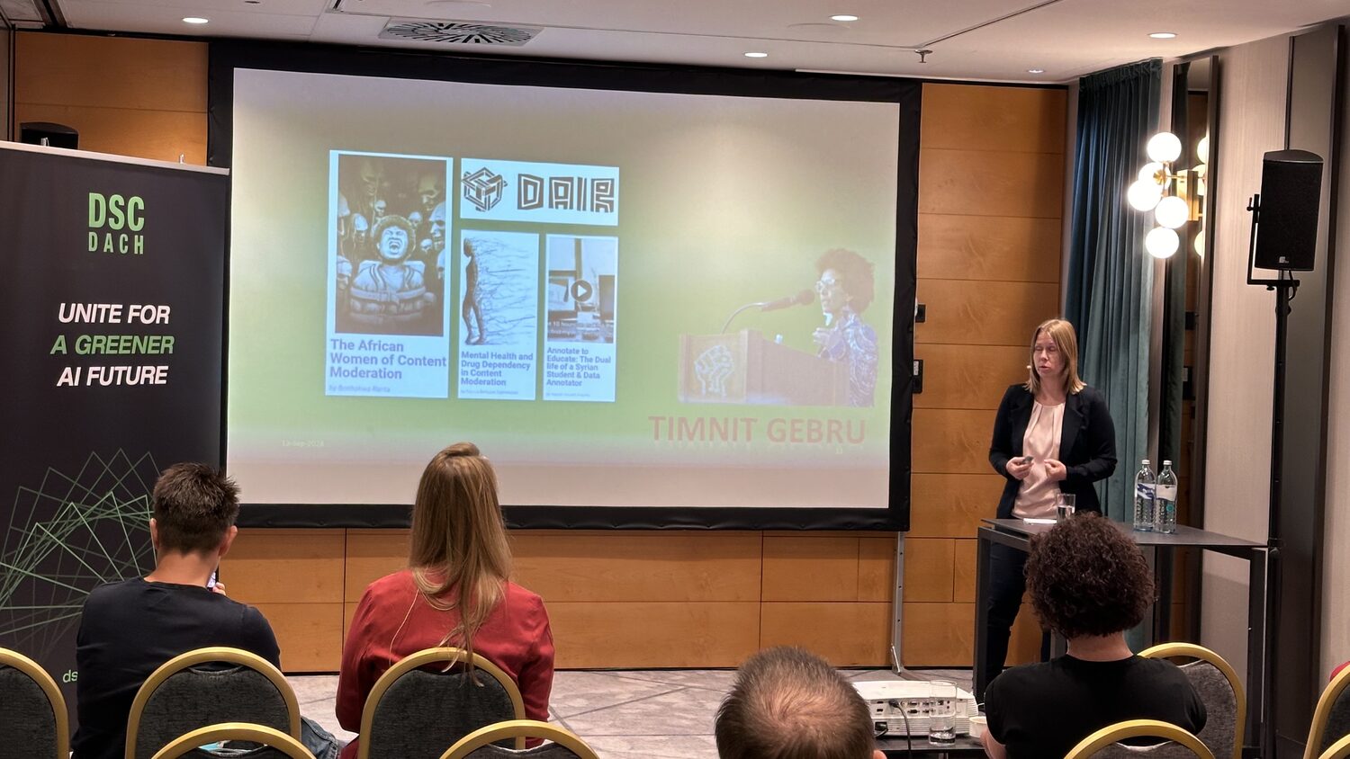 A speaker beside a slide showing the DAIR institute and Timnit Gebru, next to a DSC DACH banner reading Unite for a greener AI future.
