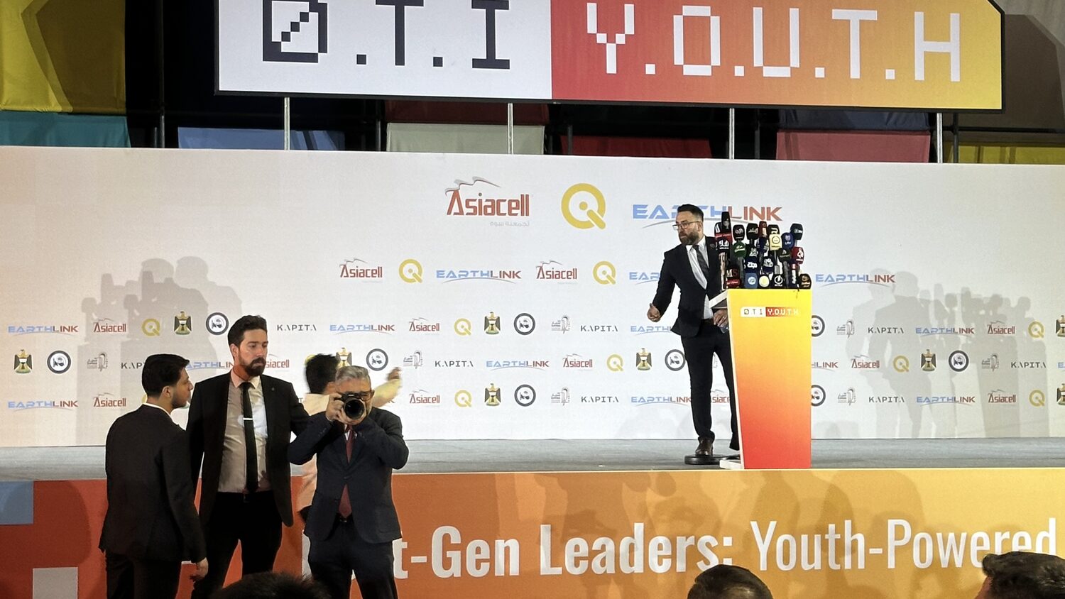 A speaker crosses to the podium at the D.T.I Y.O.U.T.H conference in Baghdad, a bank of about a dozen television microphones in front of him. Three men and a press photographer stand at stage left against a sponsor wall repeating Asiacell, EarthLink, KAPITA and the Iraqi state emblem. The overhead sign reads D.T.I Y.O.U.T.H and the stage front carries the words Youth-Powered.