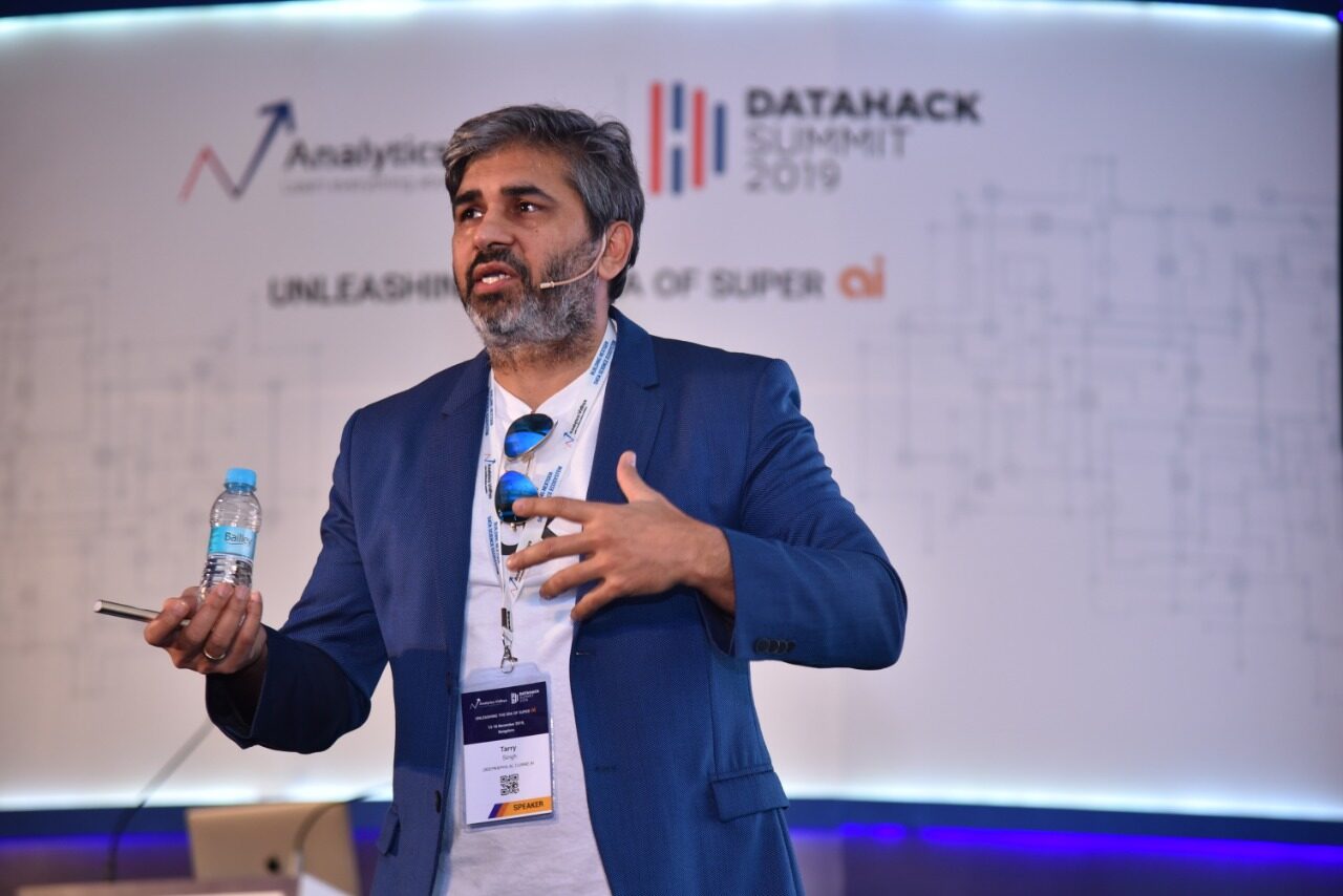 Tarry Singh speaking on the DataHack Summit 2019 stage in Bengaluru, headset microphone on, the Analytics Vidhya and DataHack Summit 2019 backdrop behind him.