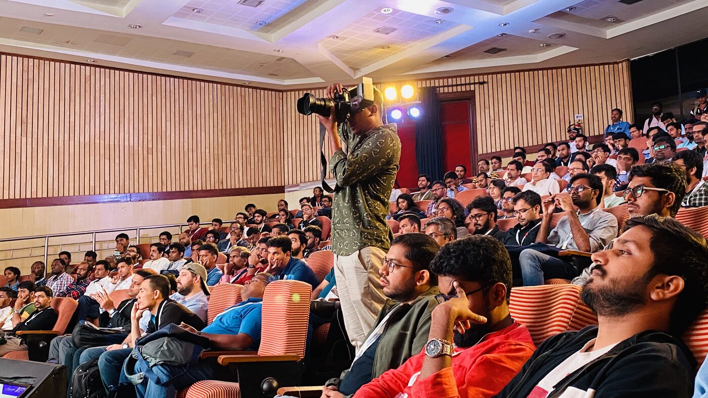 A packed auditorium of delegates during a DataHack Summit 2019 session, an event photographer standing in the centre aisle with a long lens.