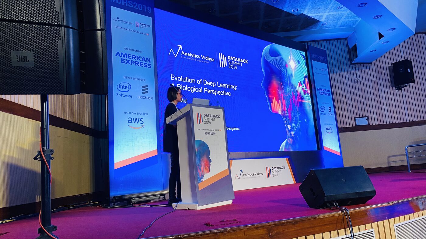 Jie Mei presenting "Evolution of Deep Learning: A Biological Perspective" from the DataHack Summit 2019 podium, the title slide on the screen behind her.