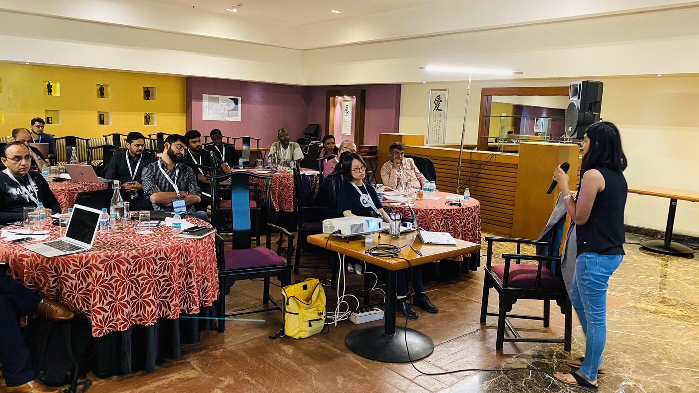 The AI Playbook workshop room at the Royal Orchid, participants seated at round tables with laptops while a woman addresses the group through a handheld microphone.