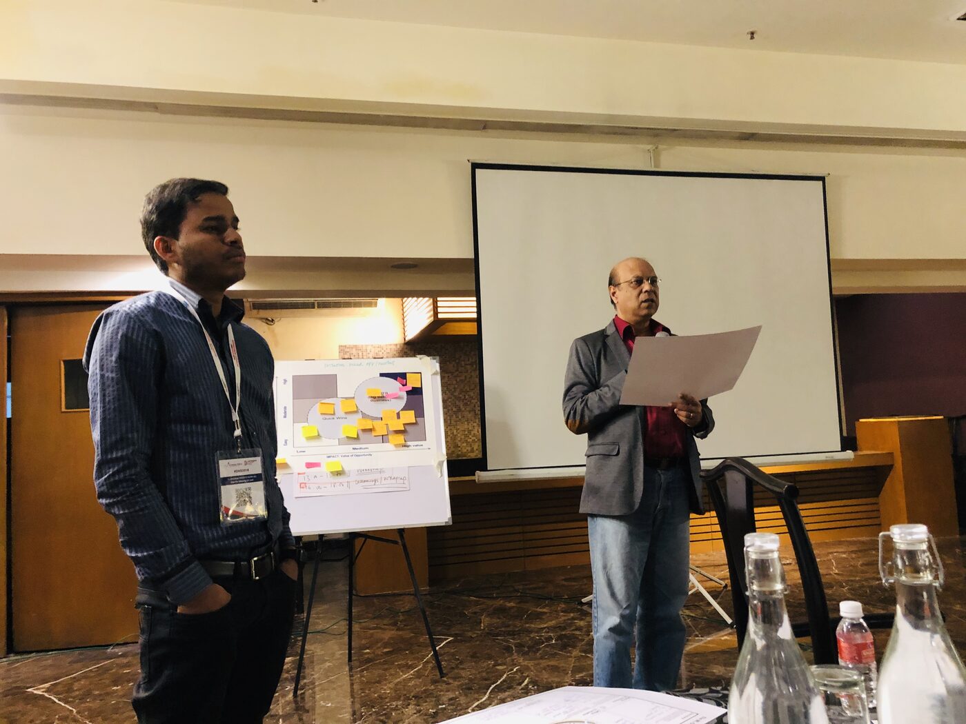Two workshop participants present their group's work to the room, standing beside a flip chart holding an impact-versus-effort prioritisation matrix covered in sticky notes.