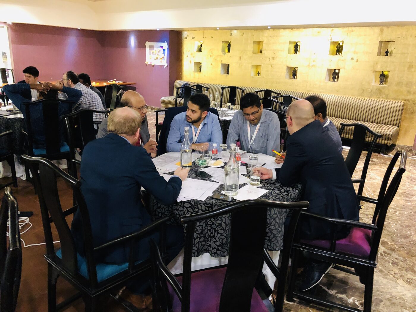 Six participants working together at a round table, marking up printed workshop canvases with pencils and coloured markers.