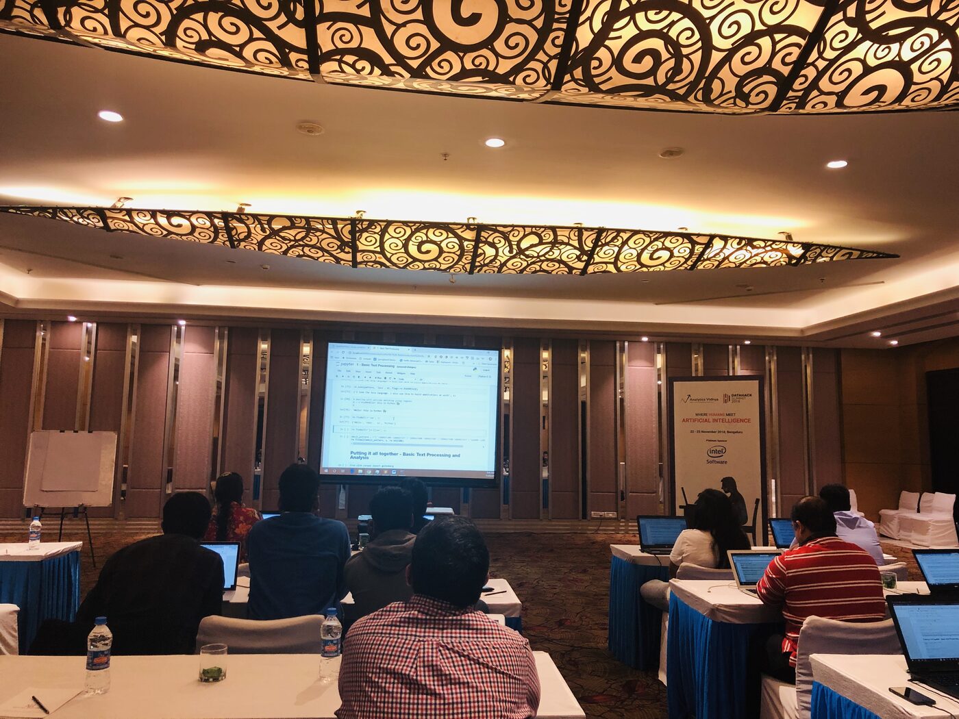 A DataHack Summit 2018 workshop room in Bengaluru: participants at laptops follow a notebook on basic text processing, beside a summit standee reading 22 to 25 November 2018, Bengaluru.