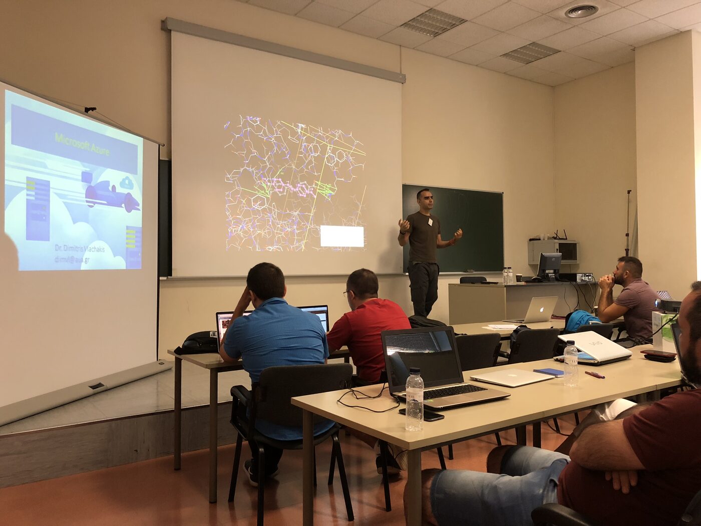 A speaker at the chalkboard at the front of the UPC classroom while participants follow on laptops, a molecular structure and an Azure title slide projected on the wall beside him.
