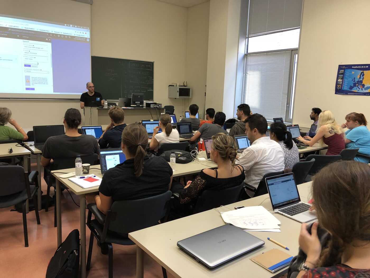 A full classroom of participants at long tables with laptops open, the instructor working the room from the back.