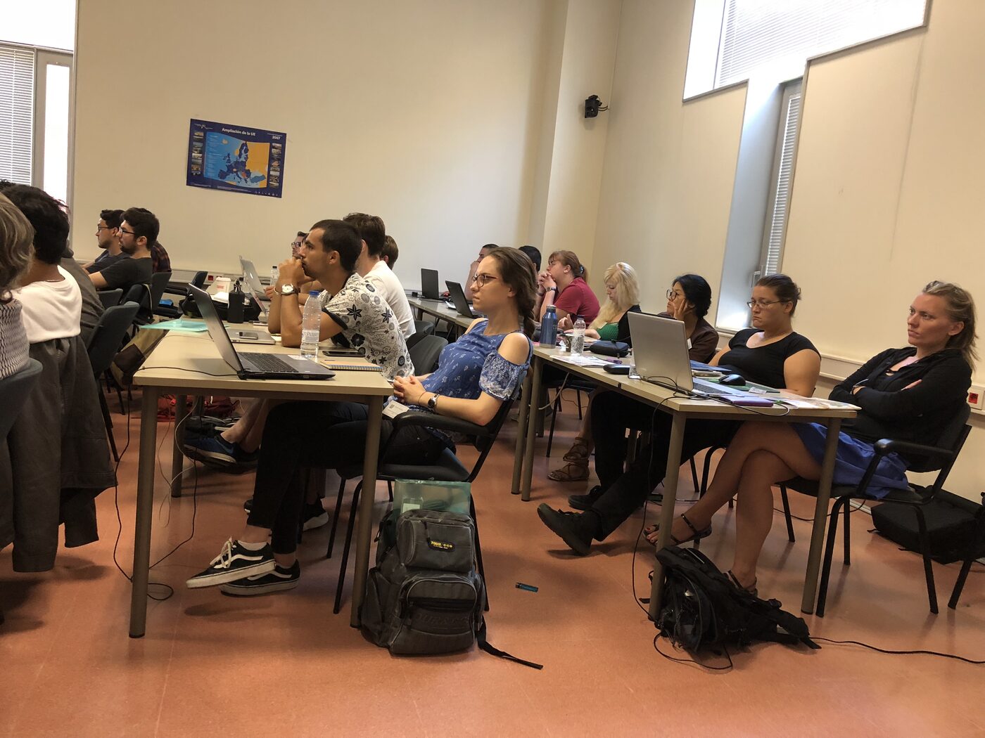 A full room of workshop participants seated at long tables with laptops, watching the front of the class.