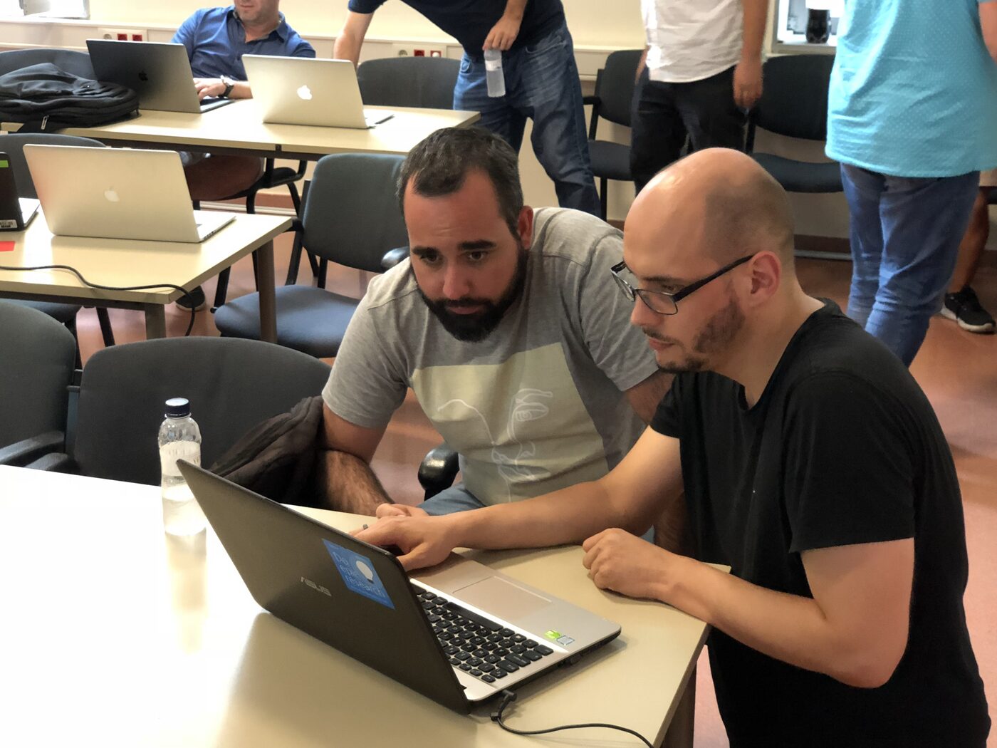 Two participants debugging code on a laptop on the workshop floor while others move around the room behind them.
