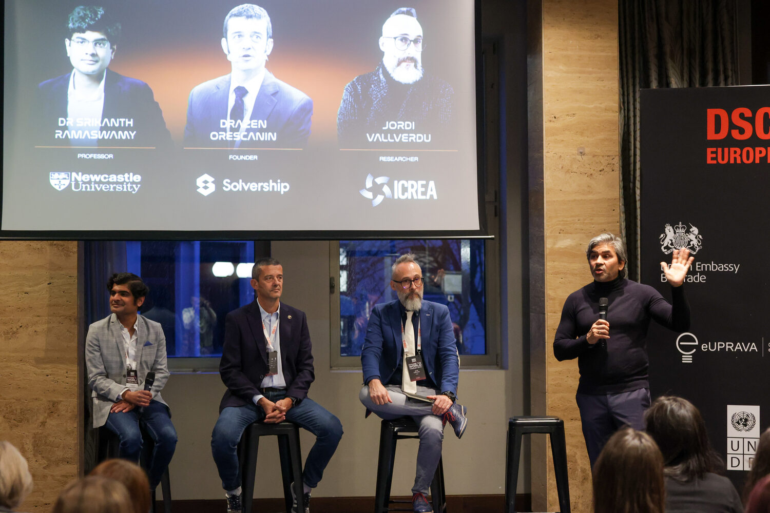 Tarry Singh stands with a microphone beside three seated panellists, the screen above naming Dr Srikanth Ramaswamy of Newcastle University, Dražen Orešćanin of Solvership and Jordi Vallverdú of ICREA, with British Embassy Belgrade and UNDP banners at the side