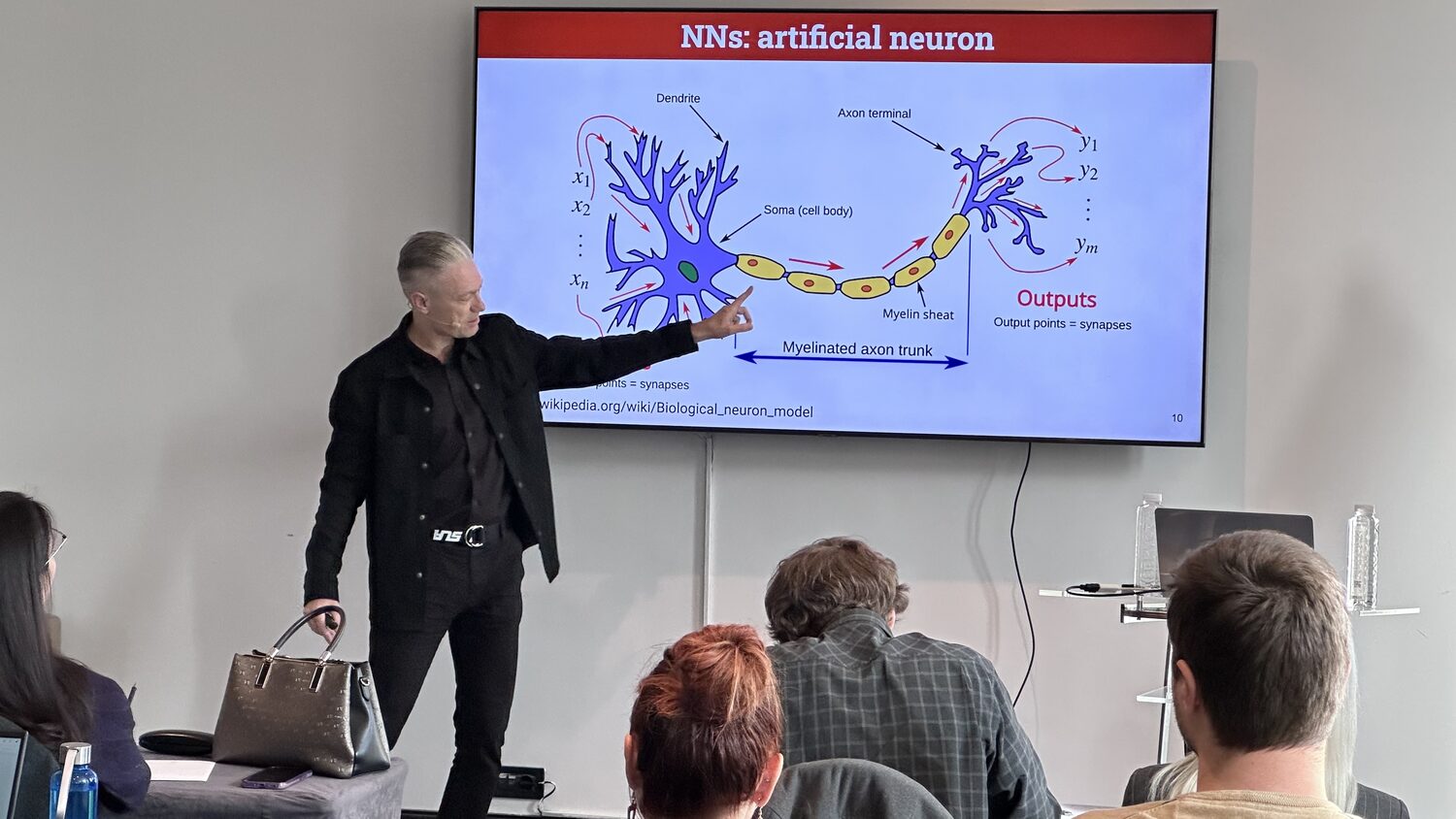 An instructor points at a screen headed NNs: artificial neuron, showing a labelled biological neuron with inputs and outputs, delegates seated in front