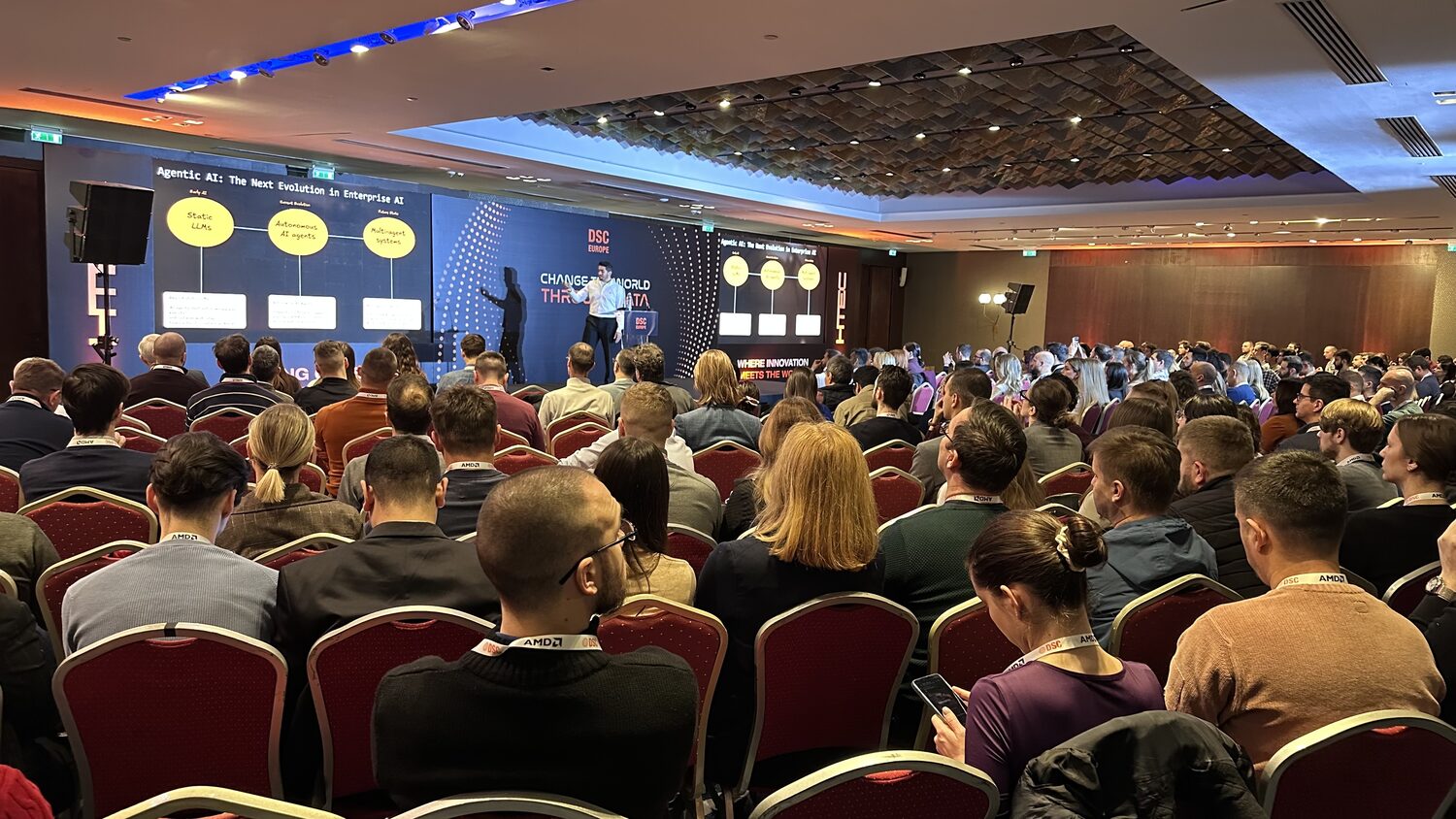 Packed main hall seen from the back, red chairs to the wall, a speaker on the DSC Europe stage beside a slide reading Agentic AI: The Next Evolution in Enterprise AI.