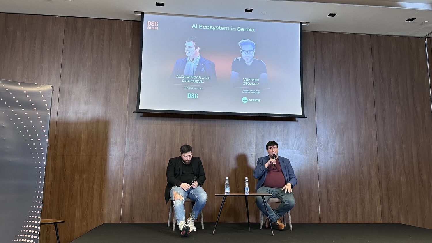 Two panellists seated on a low stage under a screen titled AI Ecosystem in Serbia, with headshots of Aleksandar Linc Djordjevic of DSC and Vukasin Stojkov of Startit.