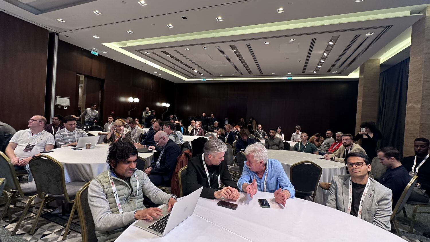 Roundtable room filling up, delegates at white-clothed round tables with laptops and lanyards, wood-panelled walls behind.