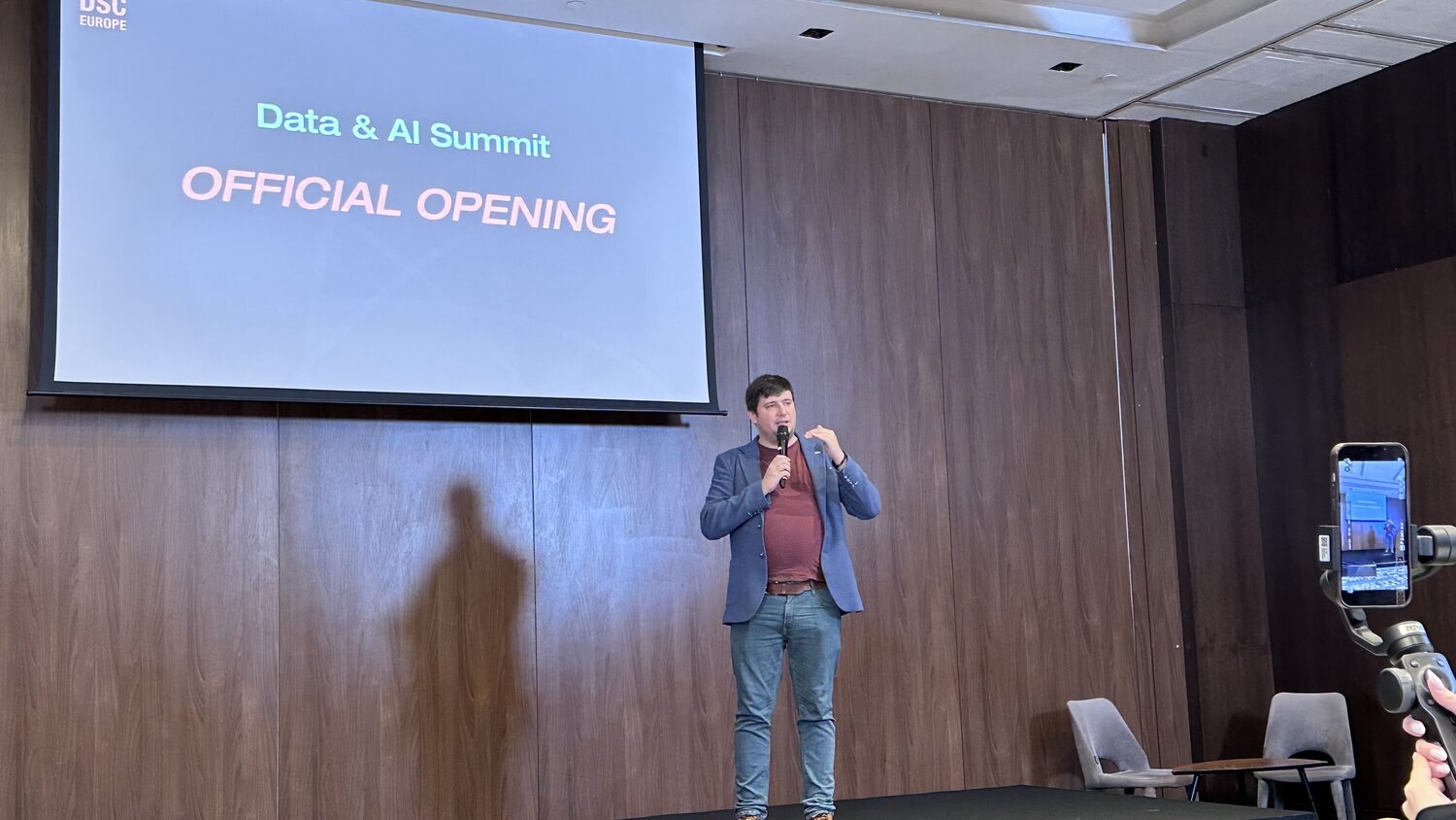 Speaker with a handheld microphone in front of a screen reading Data & AI Summit, Official Opening, under the DSC Europe mark.