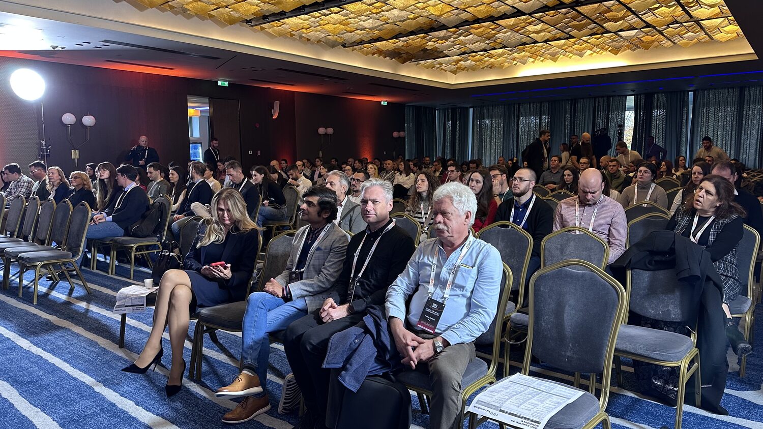 Seated audience under a gold coffered ceiling, delegates in the front row, a printed DSC Europe 25 schedule for 19 November left on a chair.