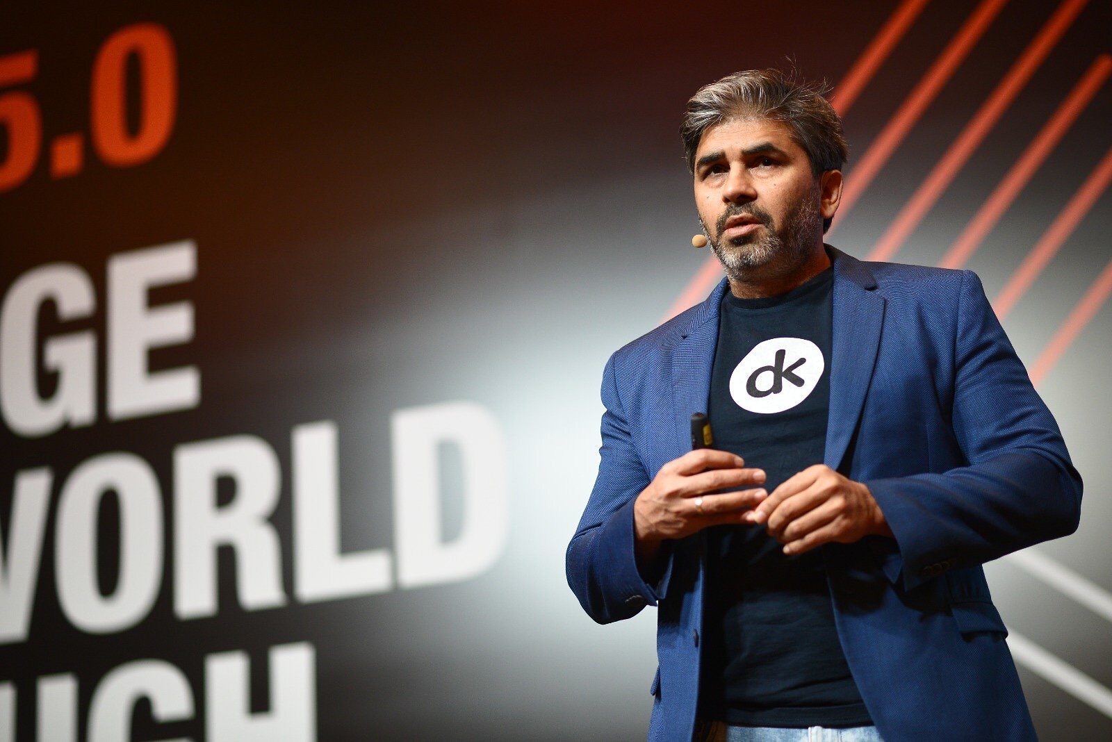 Tarry Singh presenting in a dk T-shirt and jacket, lapel microphone on, against the CHANGE THE WORLD THROUGH DATA backdrop.