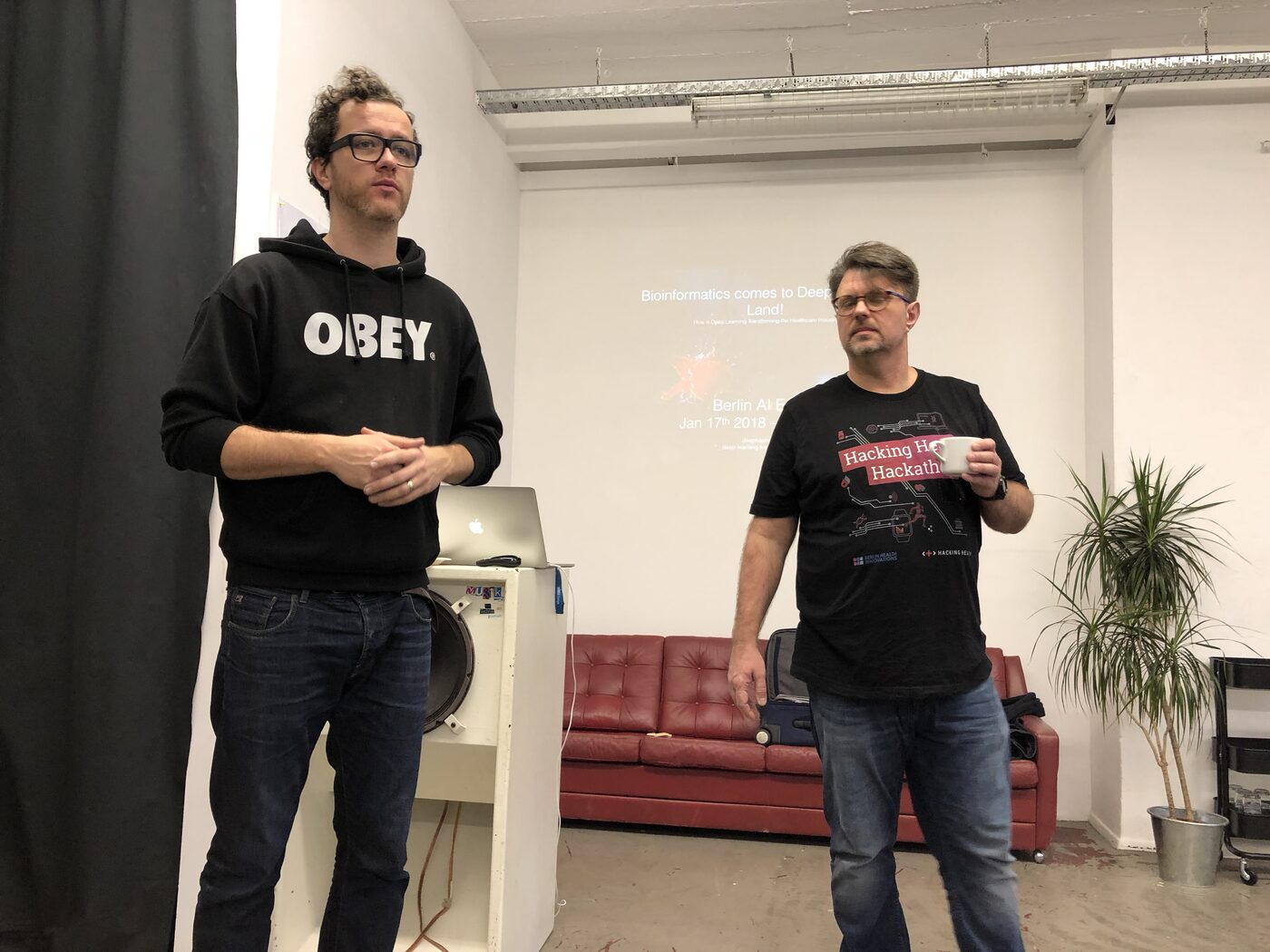 Two men presenting at the front of a Berlin workspace, a laptop on a plinth beside them and the deepkapha.ai title slide projected on the wall behind, reading "Bioinformatics comes to Deep Learning Land! How is Deep Learning Transforming the Healthcare Industry", Berlin AI, 17 January 2018.