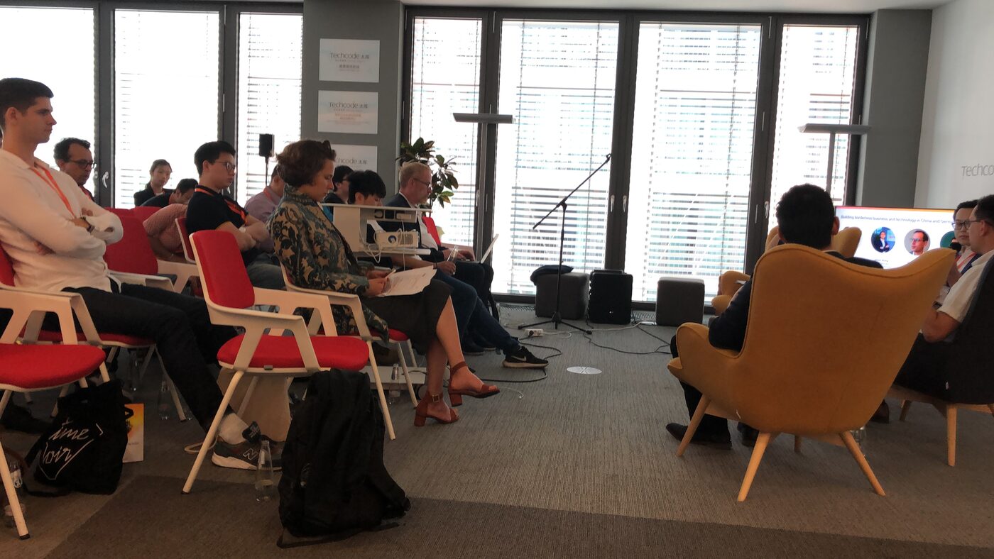 Audience seated in rows at the GCAAI annual meeting in Techcode's Berlin office, facing panellists in armchairs and a screen reading "Building borderless business and technology in China and Germany", with Techcode signage on the glass partitions.