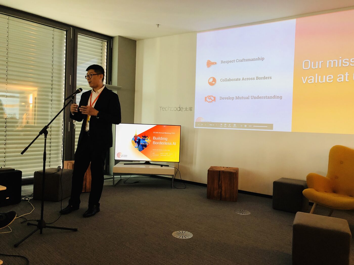 A speaker at a microphone stand in the Techcode Berlin room, beside a monitor showing the GCAAI Annual Meeting 2019 title slide, "Building Borderless AI", dated 15.06.2019, with the association's mission points projected on the wall behind.