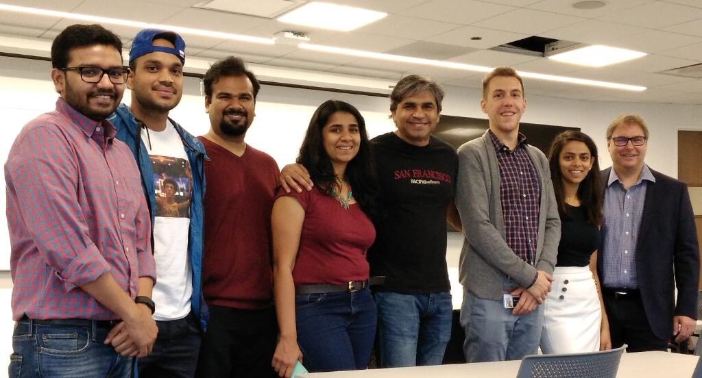 Nine participants and Tarry Singh grouped together in the teaching room, whiteboard and ceiling-mounted screen behind them, desks in the foreground.
