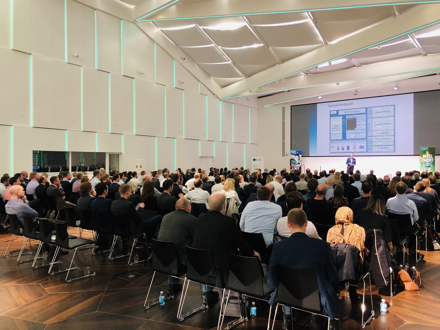 Wide view of the full hall during the Knowit programme in Copenhagen: about two hundred seated attendees on a wooden floor, a single wide projection screen, a speaker on the low stage, and green Knowit pull-up banners on both sides.