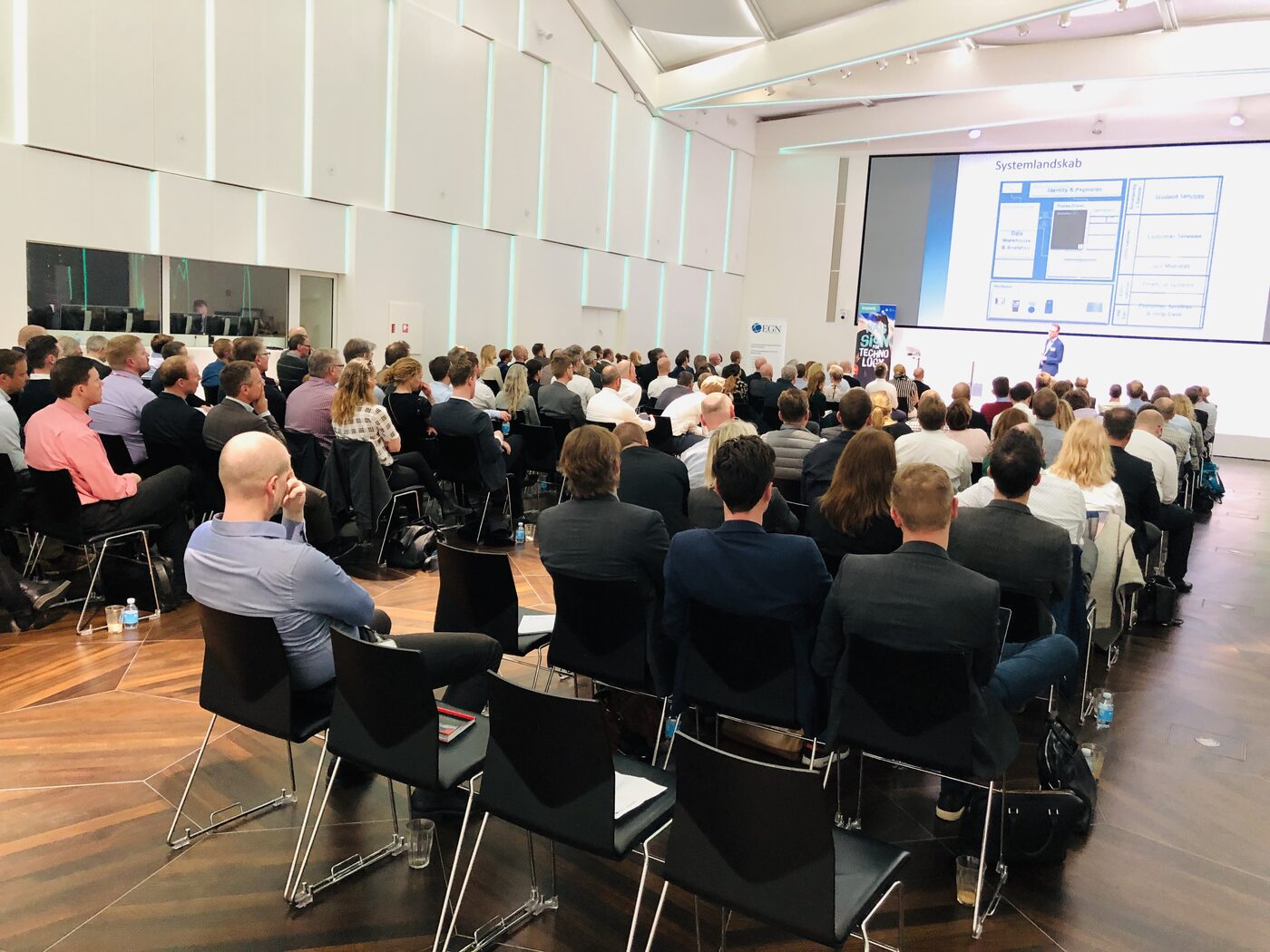 The same room seen from the back rows, attendees facing the stage while a speaker presents a slide titled Systemlandskab, with a Knowit banner standing beside the platform.
