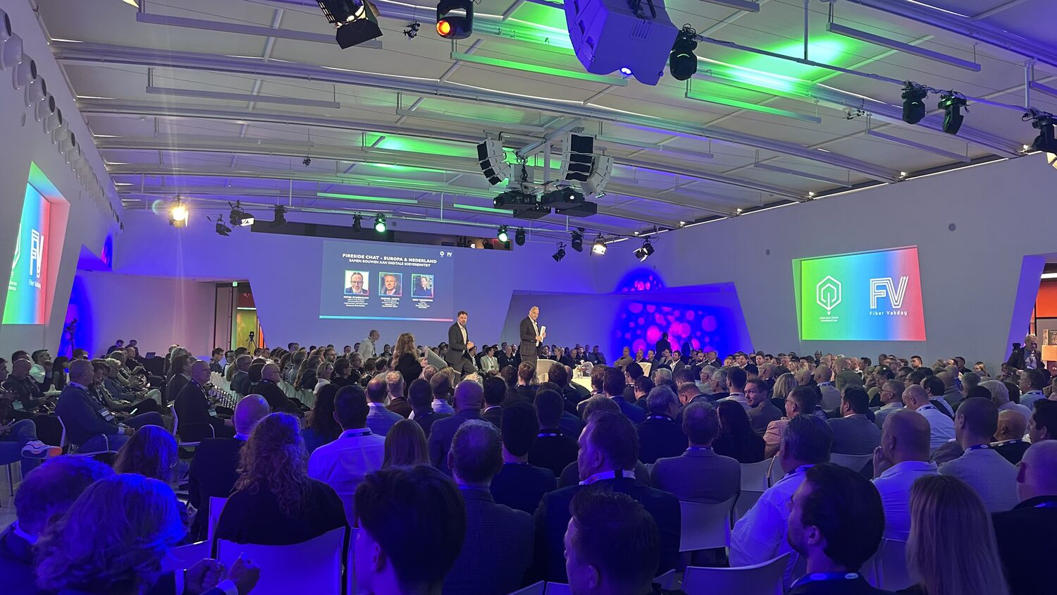 A full plenary hall at Jaarbeurs Utrecht, two speakers on a low white stage, the fireside chat panel card on the screen behind and the Fiber Vakdag mark projected on the side wall.