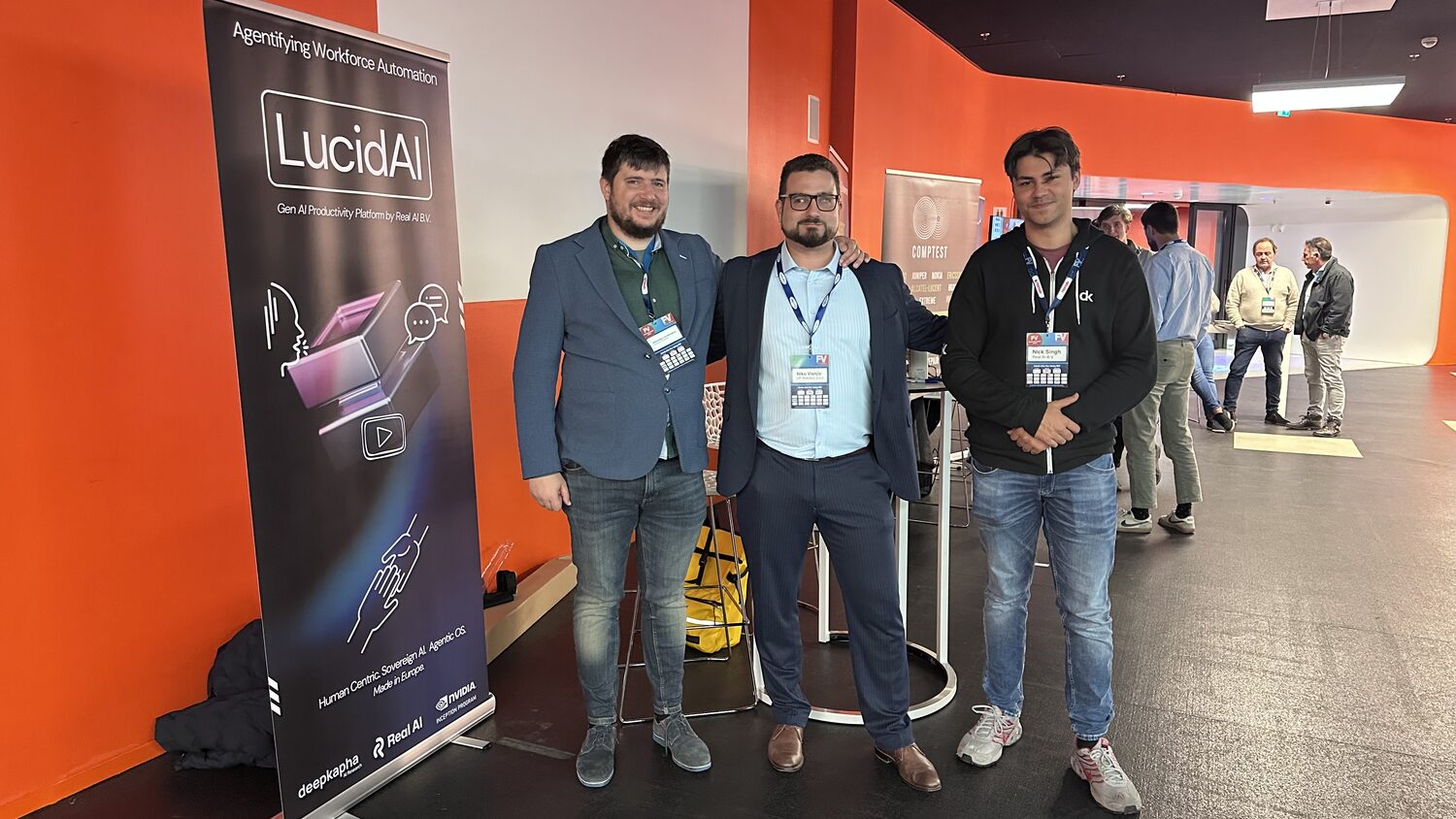 Three delegates standing beside the LucidAI roll-up banner on the exhibition floor, the Comptest stand behind them.