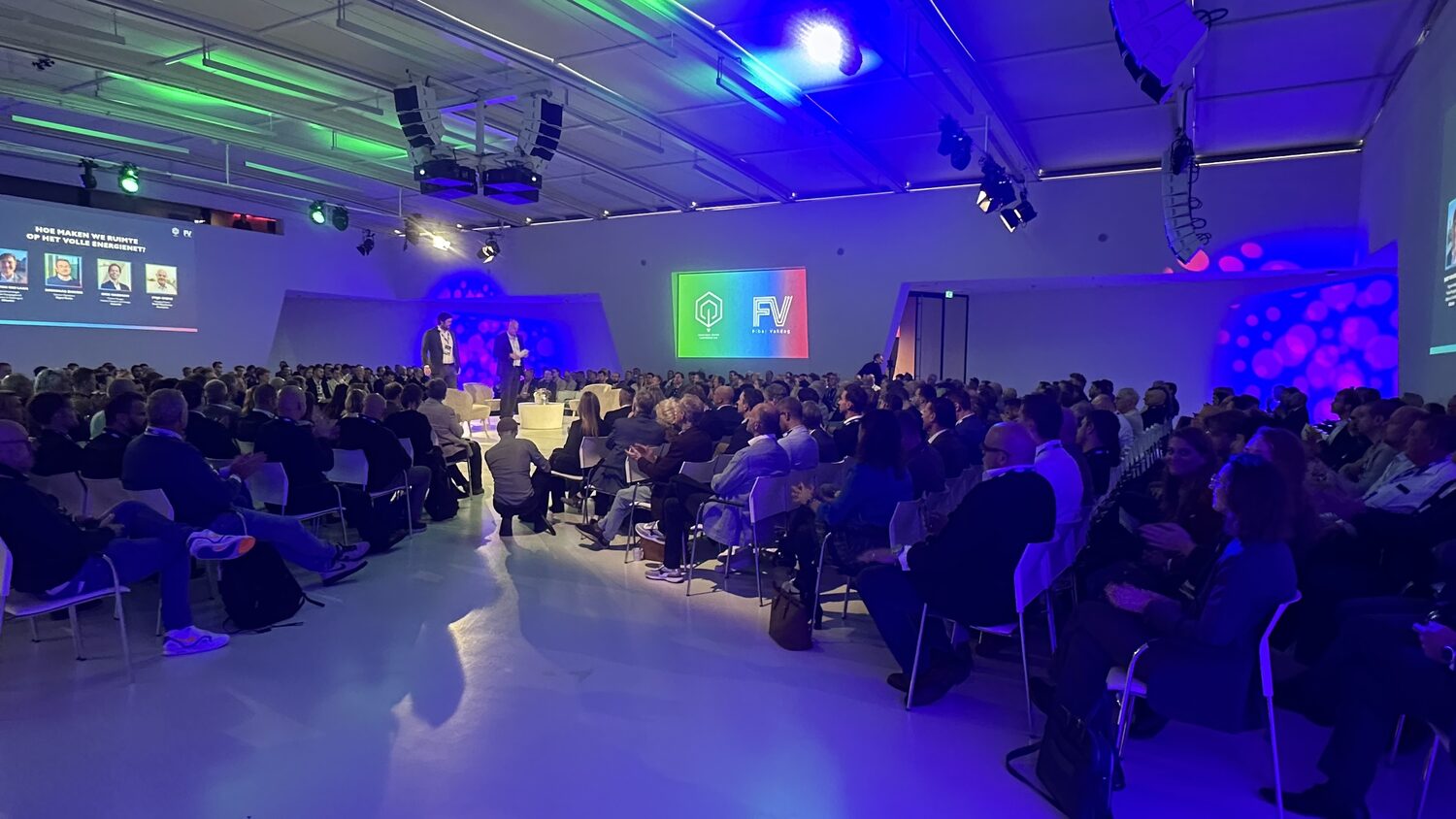 Wide view of the plenary from the back, delegates seated in rows, two speakers on stage and the Fiber Vakdag logo on the far screen.