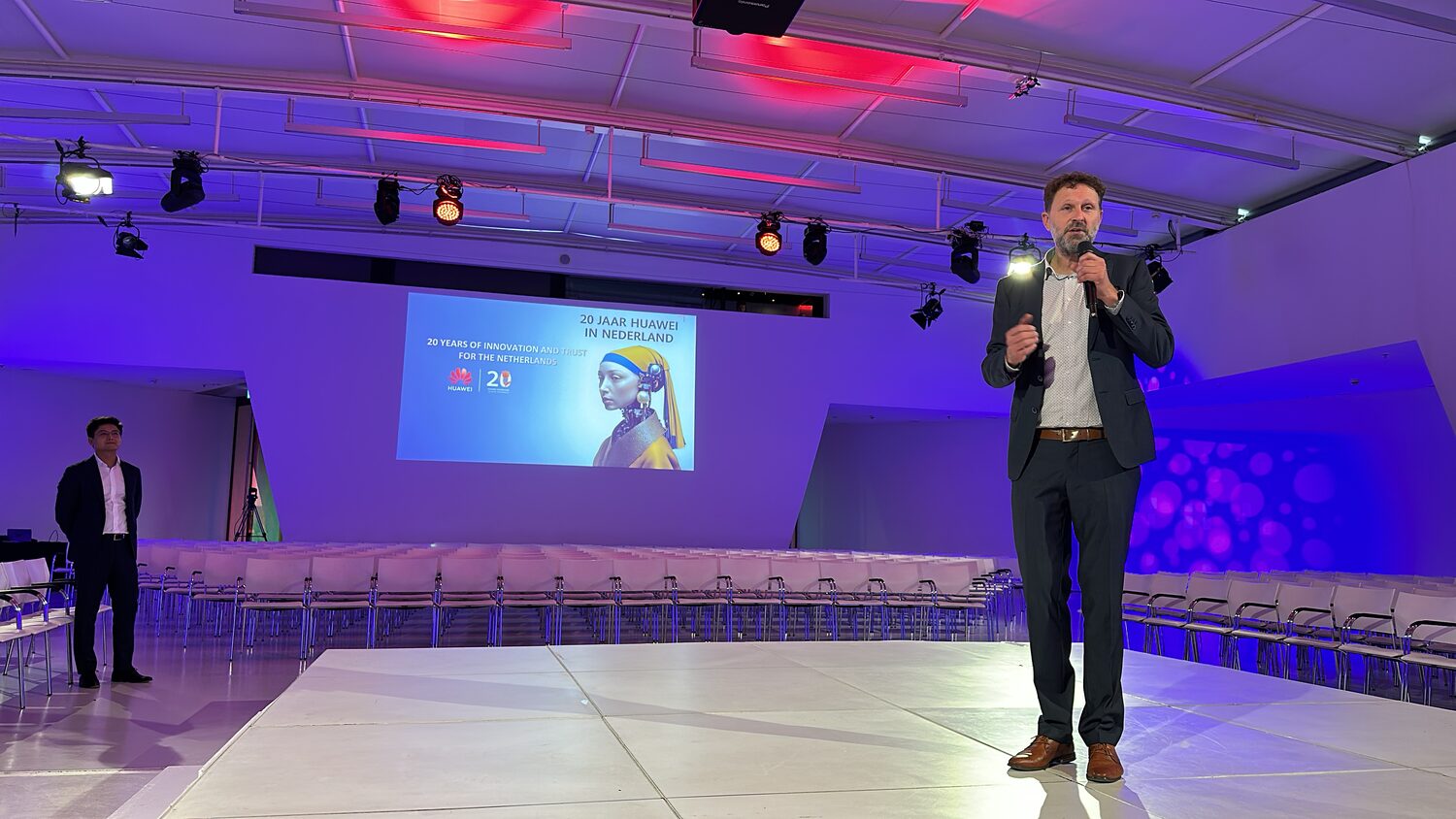 A speaker crossing the plenary stage under blue and red lighting, the screen behind reading twenty years of Huawei in the Netherlands.