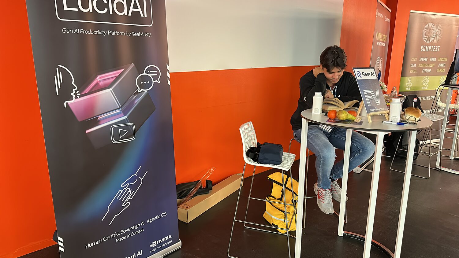 The Real AI stand, a LucidAI roll-up banner to the left and a high table carrying a Fiber Vakdag 2025 card, with the Comptest stand beyond.