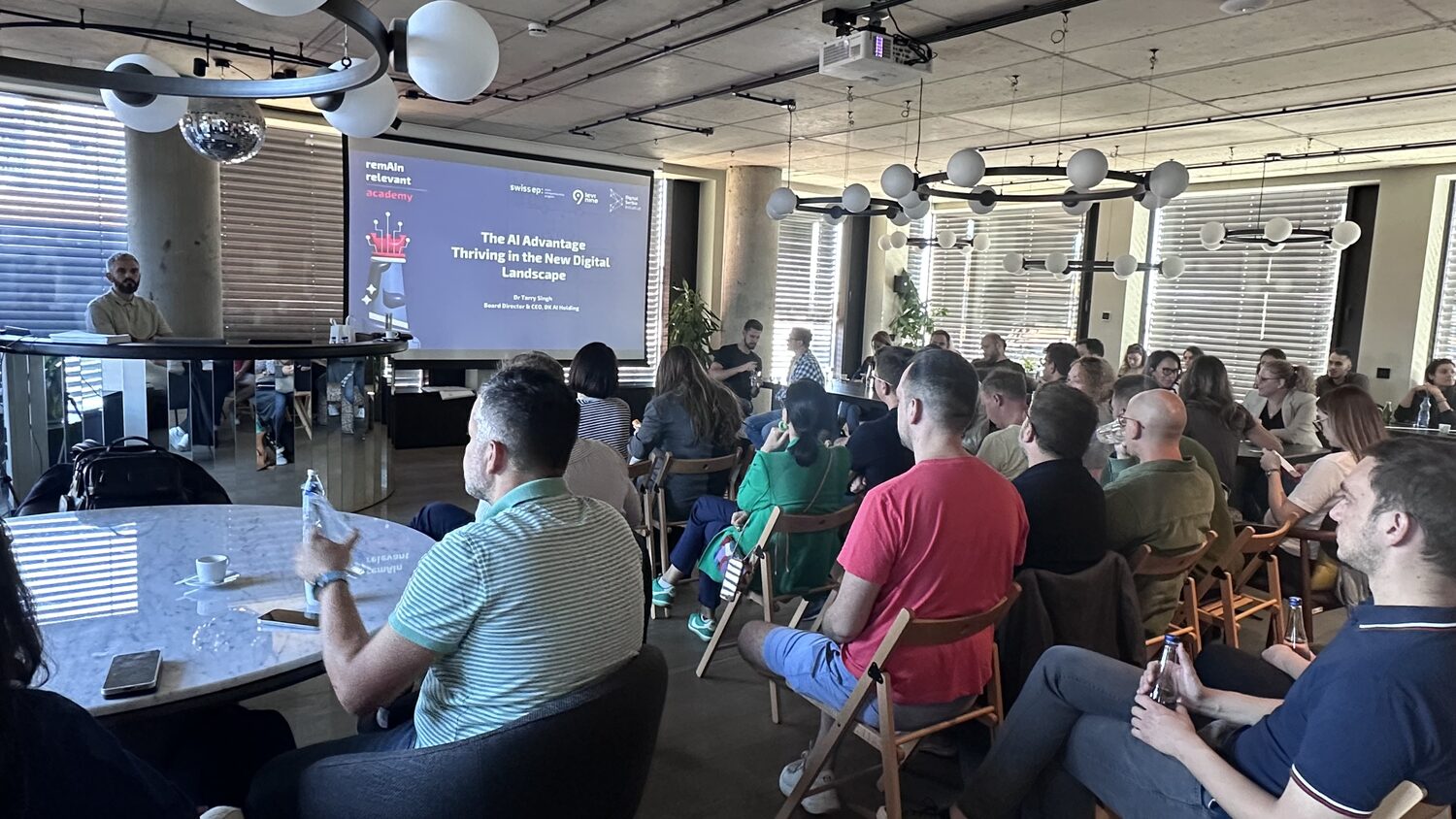 Wide view of the workshop room, rows of folding chairs facing a projection screen reading The AI Advantage.