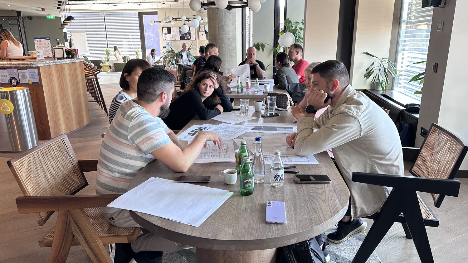 Six people around a wooden table with printed worksheets, water bottles and phones, more groups working behind them.