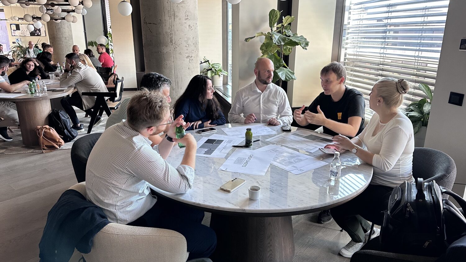 A breakout group of seven at a round table by the window, papers spread between them.