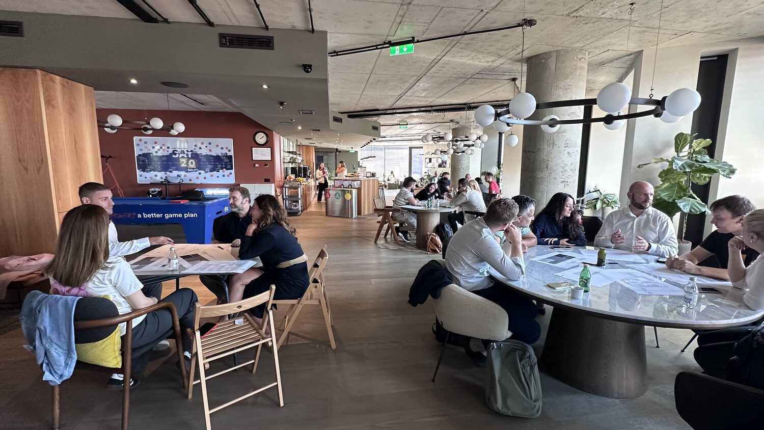 Open office floor with several breakout groups working at separate tables, a table football table against the far wall.