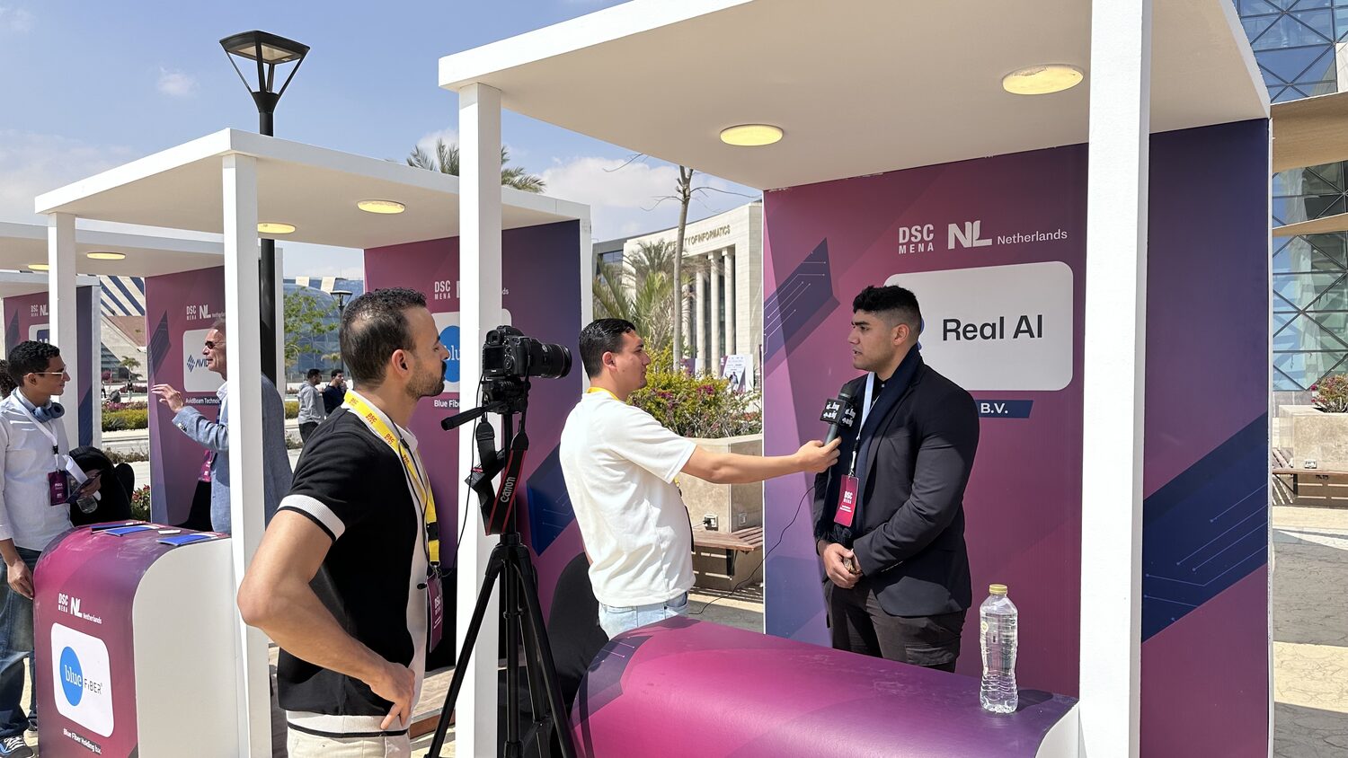 A television interview at the Real AI stand at DSC MENA 2025, with the Egypt University of Informatics building behind the row of white pavilions.