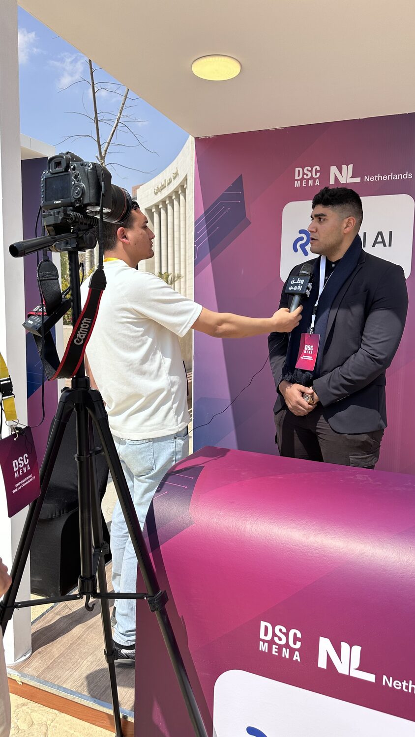 A camera operator and a reporter recording an interview against the Real AI backdrop at the DSC MENA stands.