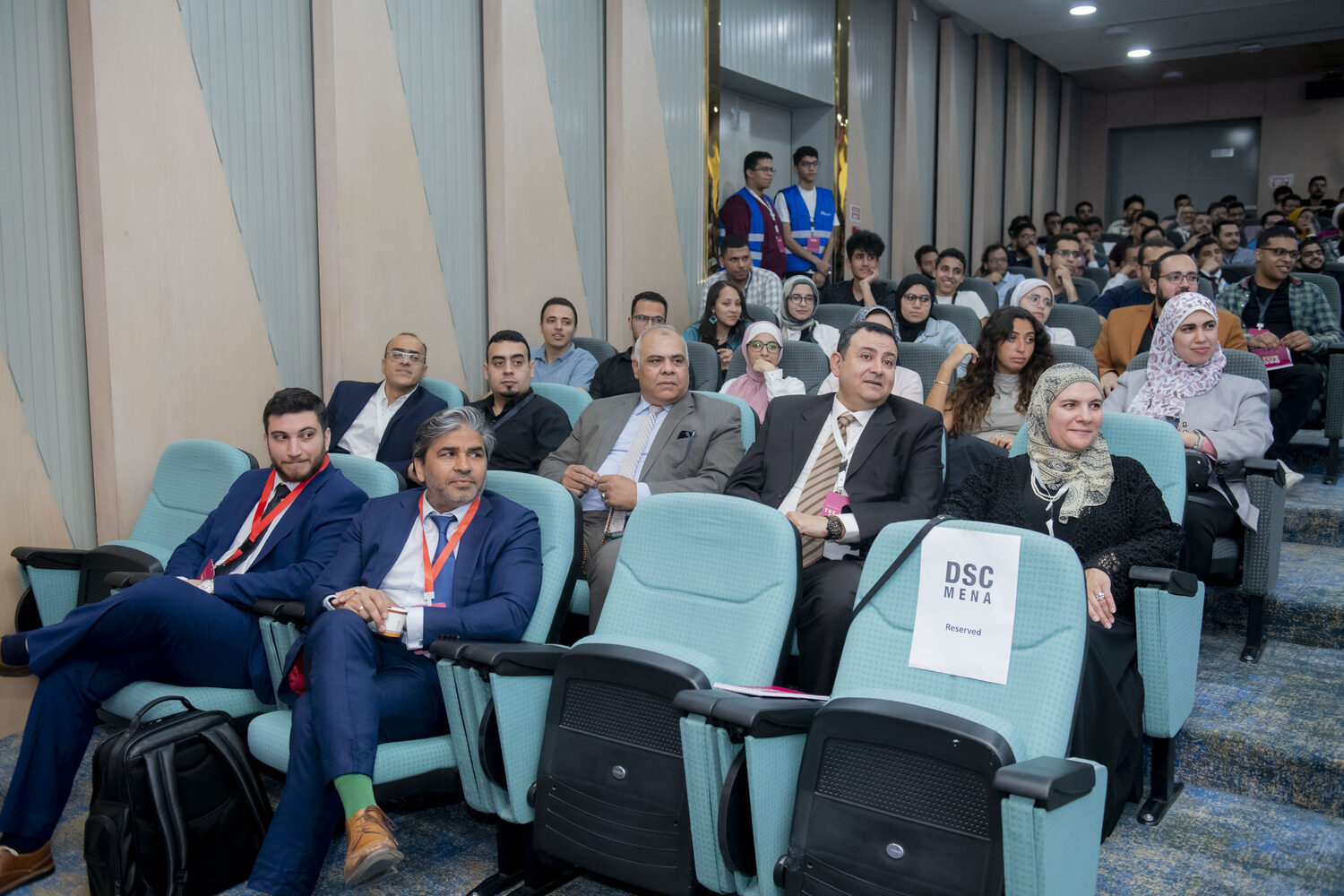 Delegates seated in the university auditorium, a DSC MENA reserved card on a front-row seat and volunteers in blue vests at the door.